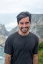 A smiling man in a black shirt poses in front of a scenic coastal cliff in Portugal.