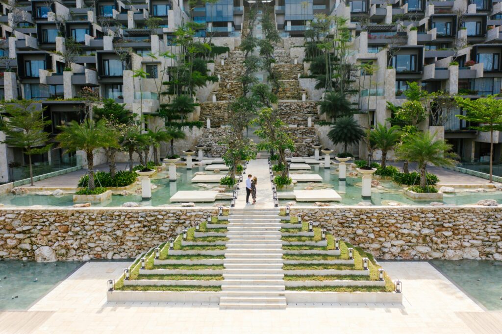 A couple walks through a modern resort with lush gardens and water features, showcasing luxurious architecture.