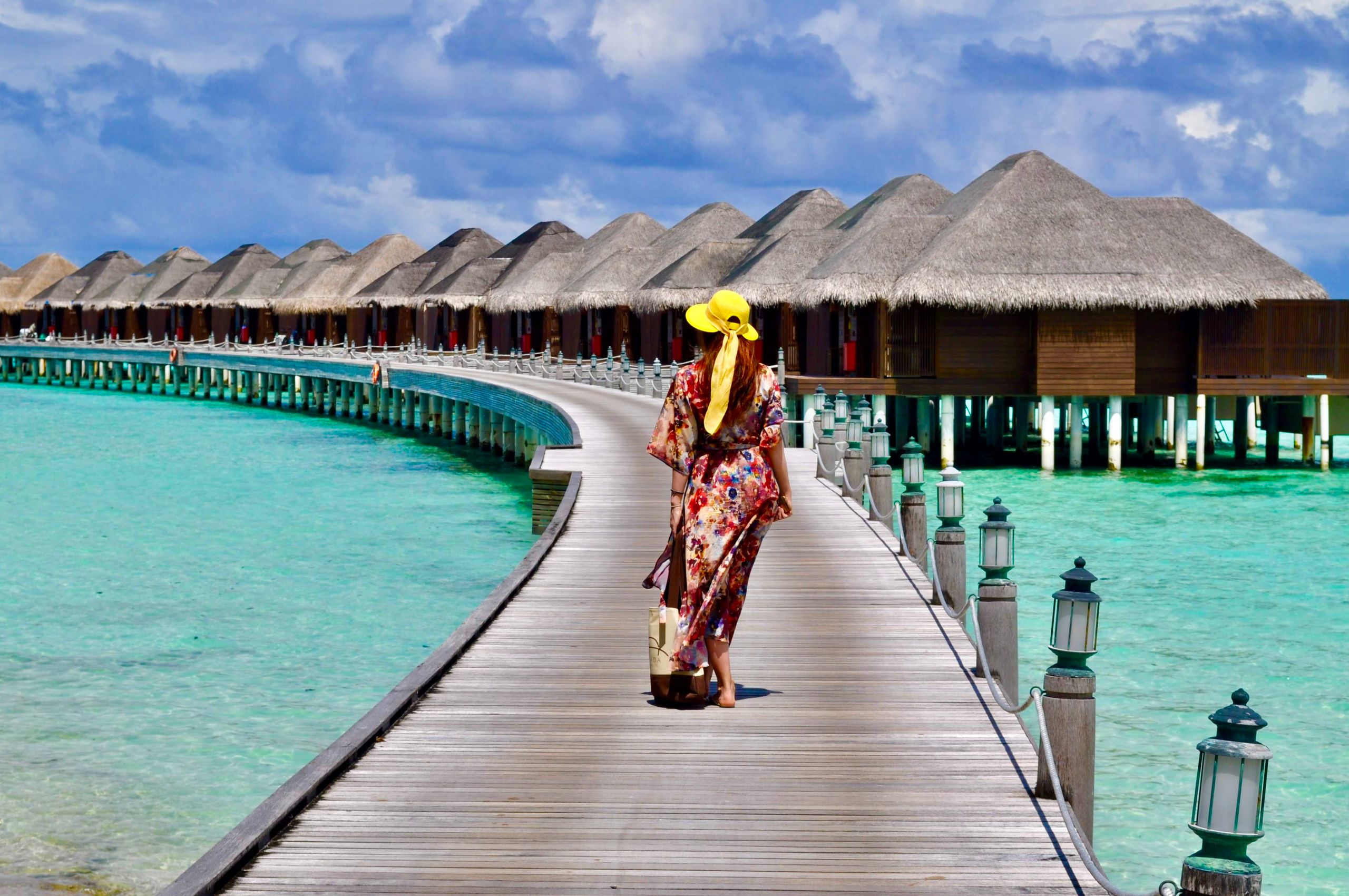 Elegant woman in summer attire walking on a boardwalk toward stunning overwater villas in the Maldives.